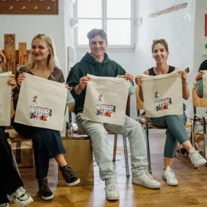 Pet nasmejanih mladih sedi v vrsti. V rokah držijo krpe s poslikavo gradu.