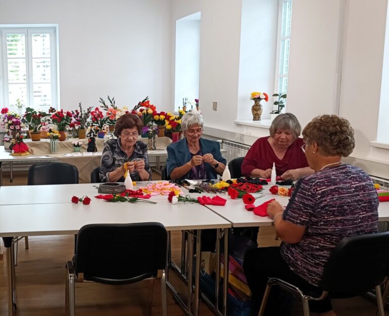 Štiri ustvarjalke sedijo ob mizi in izdeluje rože iz krep papirja. Na mizi pred njimi so že izdelane rože in dodatki za ustvarjanje. Izdelane rože so razstavljene tudi na mizi za njimi.