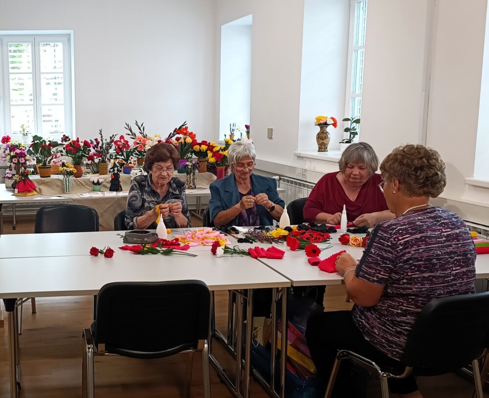 Štiri ustvarjalke sedijo ob mizi in izdeluje rože iz krep papirja. Na mizi pred njimi so že izdelane rože in dodatki za ustvarjanje. Izdelane rože so razstavljene tudi na mizi za njimi.