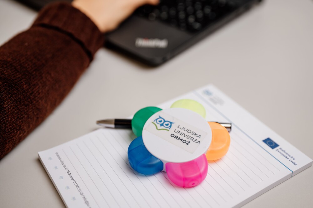 Na mizi je beležka, kuli in barvni označevalec besedila z logotipom Ljudske univerze Ormož.