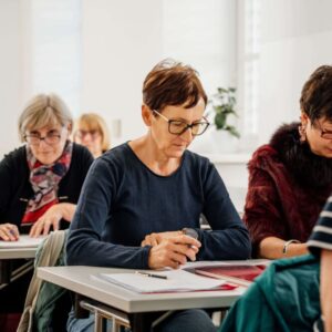 Na sliki so ljudje, ki sedijo v učilnici za mizami in se učijo.