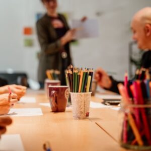 Ljudje sedijo okrog miz, na fotografiji so zamegljeni, izpostavljeni so lončki z barvicami na sredini mize.