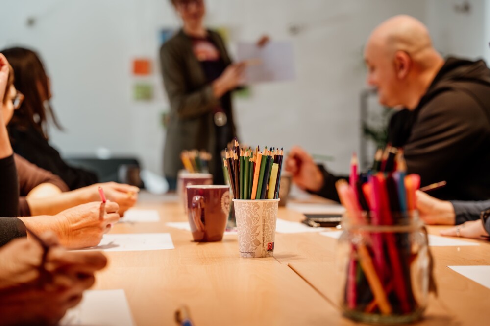 Ljudje sedijo okrog miz, na fotografiji so zamegljeni, izpostavljeni so lončki z barvicami na sredini mize.