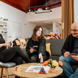 Tri osebe sedijo okrog manjše mize in igrajo igro s kartami.