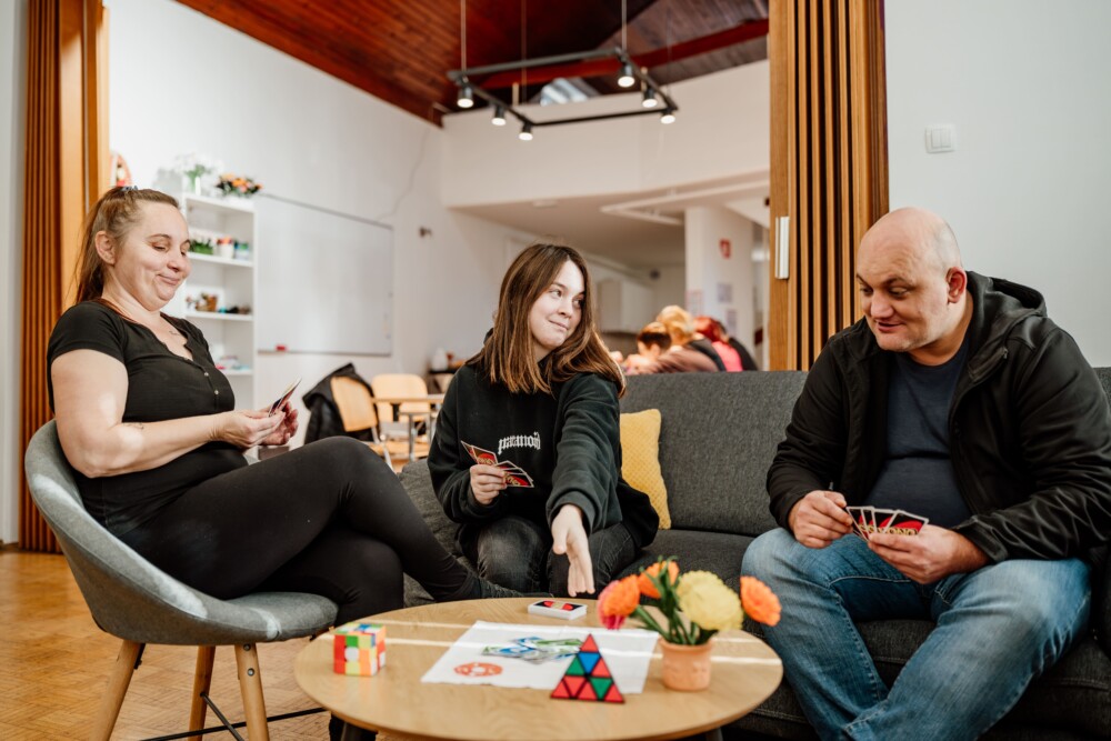 Tri osebe sedijo okrog manjše mize in igrajo igro s kartami.