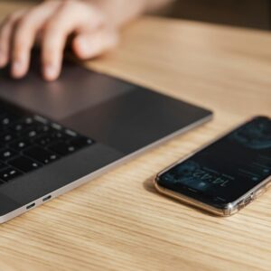 Na mizi je računalnik, na katerega tipka moški. Na desni strani poleg njega je telefon.