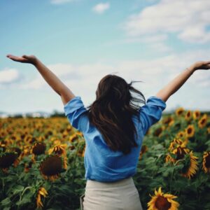 Polje sončnic, v polju stoji ženska obrnjena v drugo stran in ima roke v zraku.