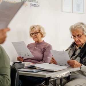Osebe sedijo v predavalnici poslušajo predavateljico in zraven spremljajo gradivo.