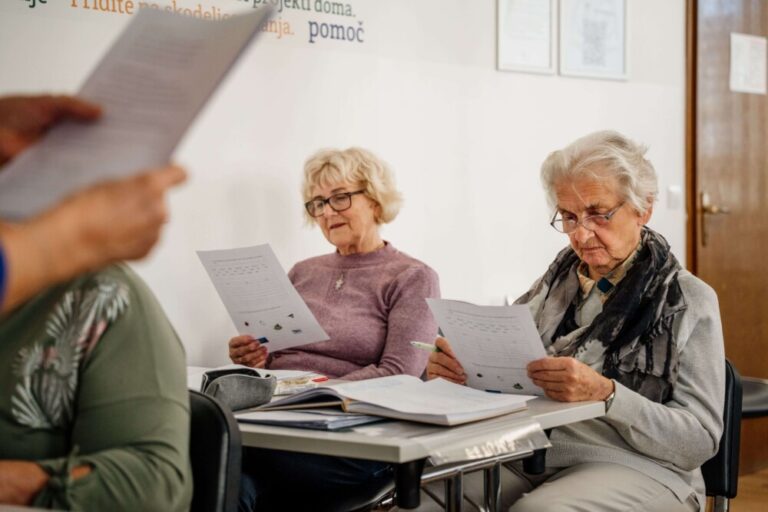 Osebe sedijo v predavalnici poslušajo predavateljico in zraven spremljajo gradivo.