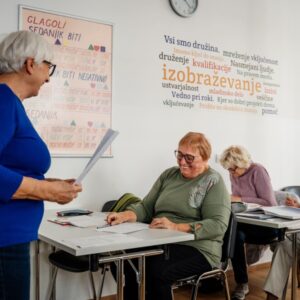 Osebe sedijo v predavalnici in poslušajo predavateljico.